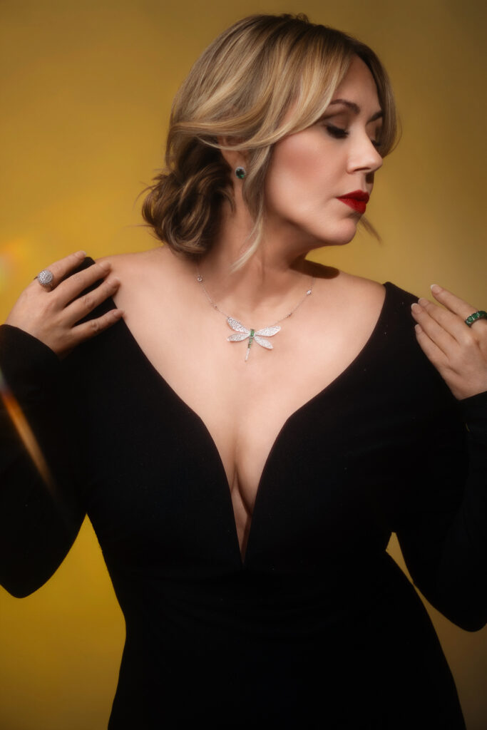 Editorial jewelry branding portrait of Darla wearing a diamond dragonfly necklace and black dress against a golden background.