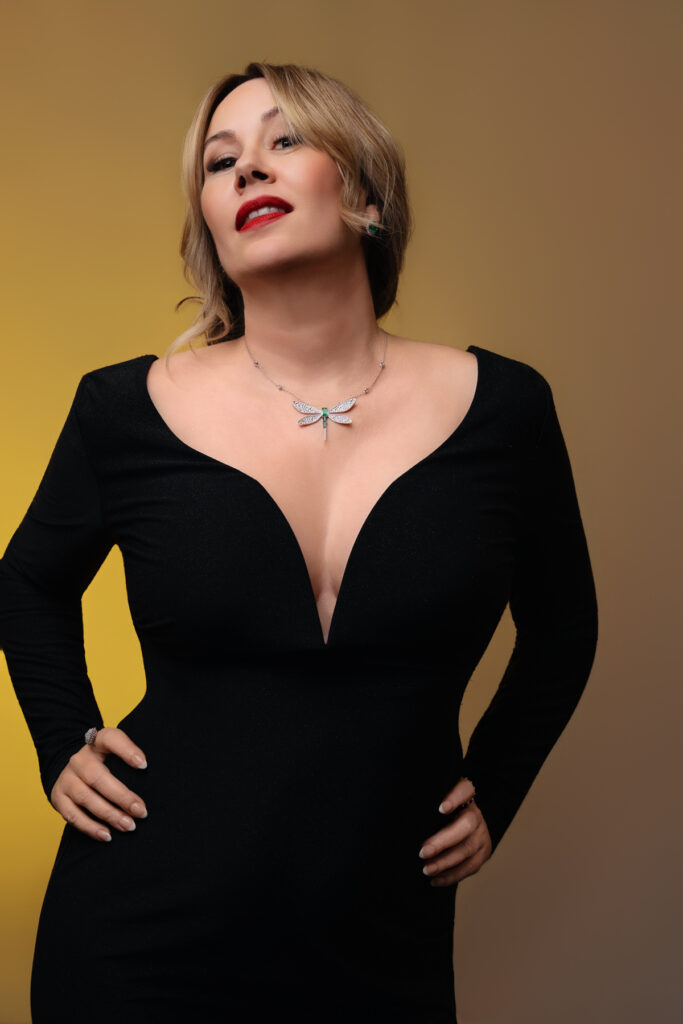 Confident entrepreneur Darla, founder of Emerald Street Designs, posing in a black dress with a dragonfly necklace against a golden backdrop.